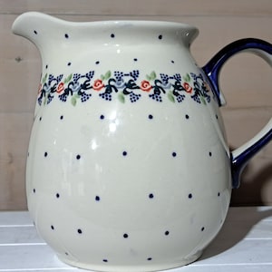 Puede incluir: Una jarra de cerámica blanca con asa azul y un diseño floral. La jarra tiene un patrón de lunares y un diseño floral azul y verde alrededor del centro.