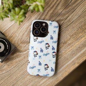 May include: A white phone case featuring a pattern of the Los Angeles Dodgers logo, blue bows, and a cartoon character wearing a Dodgers baseball cap and holding a baseball. The case is on a wooden surface.