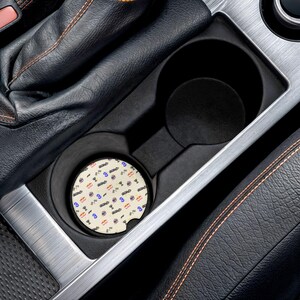 May include: A circular car cup holder coaster featuring a repeating pattern of small Formula 1 race cars, the number 3 in blue, and red bull logos on a cream-colored background, placed inside a black plastic vehicle cup holder console.