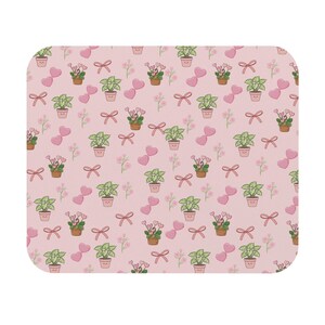 May include: A rectangular pink mousepad featuring a repeating pattern of potted plants, pink hearts, bows, and small flowers. The plants have smiling faces. The design is a cute and whimsical illustration.