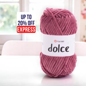 Puede incluir: Una madeja de hilo YarnArt Dolce de color rosa polvoriento. La etiqueta dice "dolce" en negrita, sin serifa. La imagen también incluye el texto "UP TO 20% OFF EXPRESS". El hilo está sobre una superficie blanca.