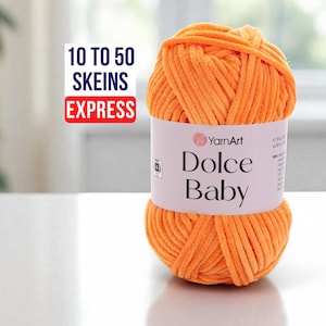 Puede incluir: Un ovillo de lana naranja YarnArt Dolce Baby. La etiqueta dice "Dolce Baby" e incluye el logotipo de YarnArt. La imagen también incluye el texto "10 a 50 madejas Express". La lana es un material suave y afelpado.