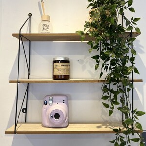 May include: A black metal shelf unit with three shelves. The top shelf has a clear glass bottle with a white label and a small plant in a pot. The middle shelf has a candle with a white label that says "Aromatherapy" and "Positive Vibes". The bottom shelf has a pink instant camera.