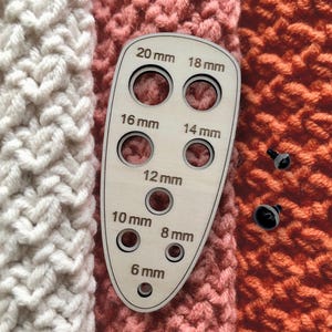 May include: A light-coloured wooden measuring tool for craft eyes with eight circular cutouts labelled 6 mm, 8 mm, 10 mm, 12 mm, 14 mm, 16 mm, 18 mm, and 20 mm. Two small black plastic safety eyes are positioned to the right of the measuring guide.