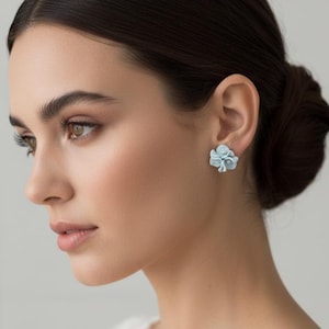 Peut inclure: Gros plan d'une personne portant des boucles d'oreilles clous en forme de fleurs bleu clair. Les boucles d'oreilles sont composées de plusieurs pétales. La personne a les cheveux foncés attachés en chignon et porte un haut en dentelle blanche.
