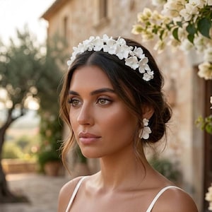 May include: A woman wearing a white floral crown and matching earrings. The crown and earrings feature delicate, three-dimensional white flowers. She is wearing a white spaghetti strap dress with lace detailing. The background includes a stone building and flowering plants.
