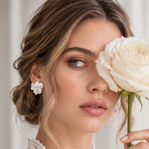 Könnte beinhalten: Ein weißer Blumen-Ohrstecker mit geschichteten Blütenblättern und einem kleinen zentralen Akzent. Der Ohrring wird am Ohrläppchen getragen und befindet sich neben einer großen, cremefarbenen Rose, die vor dem Gesicht gehalten wird.