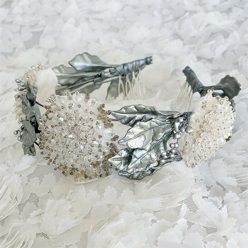 In this image we can see the bridal tiara crown with rhinestones, porcelain leaves and silver crystals on a white fabric with flowers.