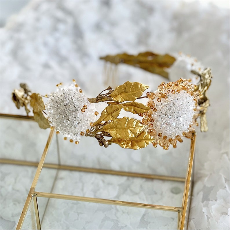 In this image we can see the bridal tiara crown with rhinestones, porcelain leaves and gold crystals on a white fabric with flowers.