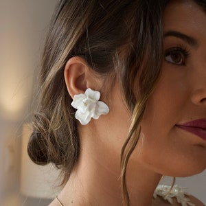 Puede incluir: Pendientes de cerámica blanca con forma de flor. Los pendientes tienen forma de flor con varios pétalos. Los pendientes son un accesorio delicado y femenino.