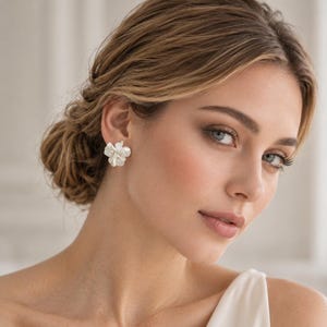 Könnte beinhalten: Ein Paar weiße Blumen-Ohrstecker mit geschichteten, blütenblattartigen Formen und einer glatten, matten Oberfläche. Die Ohrringe sind als kleine, zarte Blüten gestaltet, die flach am Ohrläppchen anliegen und ein schlichtes Design aufweisen.