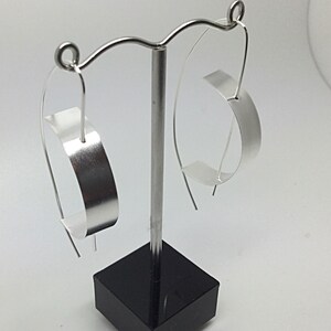 May include: A pair of silver-toned hoop earrings featuring a wide, flat, curved metal band suspended from a thin, elongated wire hook. The earrings have a smooth, reflective finish and are displayed on a stand with a black square base.