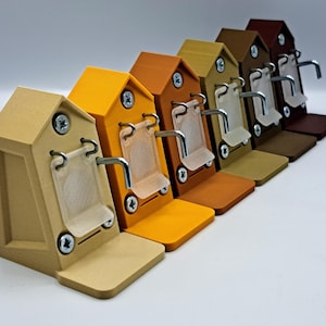 Könnte beinhalten: Eine Reihe von miniaturförmigen Schlüsselhaltern in Hausform in verschiedenen Farben wie Beige, Gelb, Orange und Braun. Jeder Halter hat einen kleinen, transparenten Kunststoffeinsatz und einen Metallhaken. Die Schlüsselhalter sind diagonal vor weißem Hintergrund angeordnet.