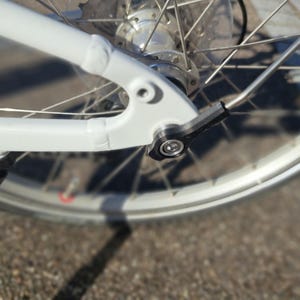 Könnte beinhalten: Nahaufnahme eines weißen Fahrradrahmens und -rads. Der Rahmen hat einen schwarzen Schnellspanner für das Hinterrad. Das Rad hat silberne Speichen und eine silberne Felge. Das Fahrrad steht auf einer strukturierten Oberfläche.