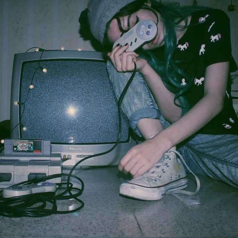 May include: A person wearing a grey beanie and a black t-shirt with white unicorns printed on it, holding a grey Super Nintendo controller. The person is sitting in front of a vintage television with static on the screen. A Super Nintendo console and game cartridge are visible in the foreground.