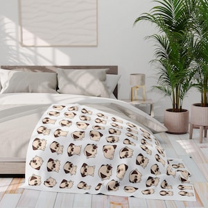 Op de afbeelding: Witte deken met een herhaald patroon van cartoonmopshonden in verschillende poses. De mopshonden zijn voornamelijk beige en bruin, met zwarte accenten. De deken is gedrapeerd over een bed met neutraal gekleurd beddengoed.