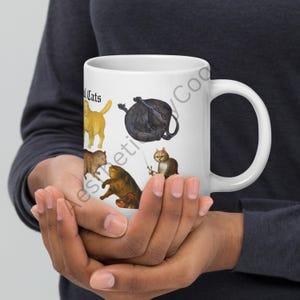May include: White ceramic mug featuring various cat illustrations in different colors and poses. The mug has the text "Medieval Cats" in a stylized font. The mug is held by a person.