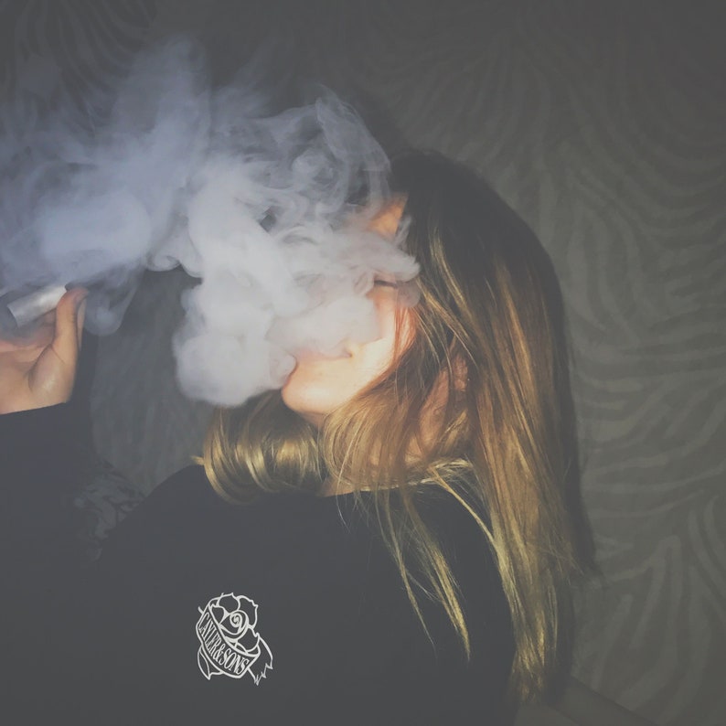 May include: A person with long blonde hair wearing a black long-sleeved shirt with a white rose and the word "Untrasonic" printed on it. The person is exhaling a large cloud of smoke.