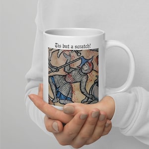 May include: White ceramic mug featuring a medieval illustration of a knight in chainmail armor, with the text "Tis but a scratch!". The artwork depicts a battle scene with a knight holding a weapon.