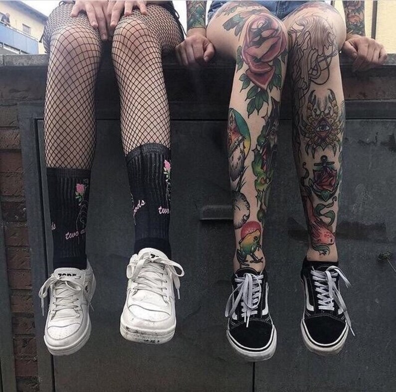 May include: Two people wearing black fishnet stockings and black socks with floral embroidery. One person has colorful tattoos on their legs. Both are wearing black and white sneakers.