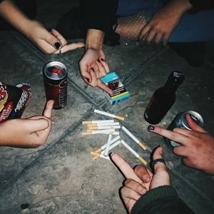 May include: A group of friends gathered around a table, sharing cigarettes, drinks, and snacks. The table is covered in a dark, textured surface. There are several cigarettes scattered on the table, along with a can of soda, a bottle of alcohol, and a bag of chips.