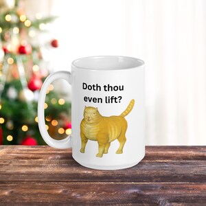 May include: White ceramic mug featuring a cartoon image of a yellow cat and the text "Doth thou even lift?" printed on it.