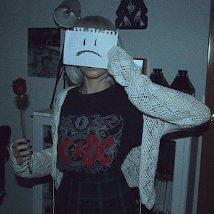 May include: A person wearing a black t-shirt with a red AC/DC logo, a white crocheted cardigan, and a plaid skirt. They are holding a single red rose and covering their face with a piece of paper with a sad face drawn on it.