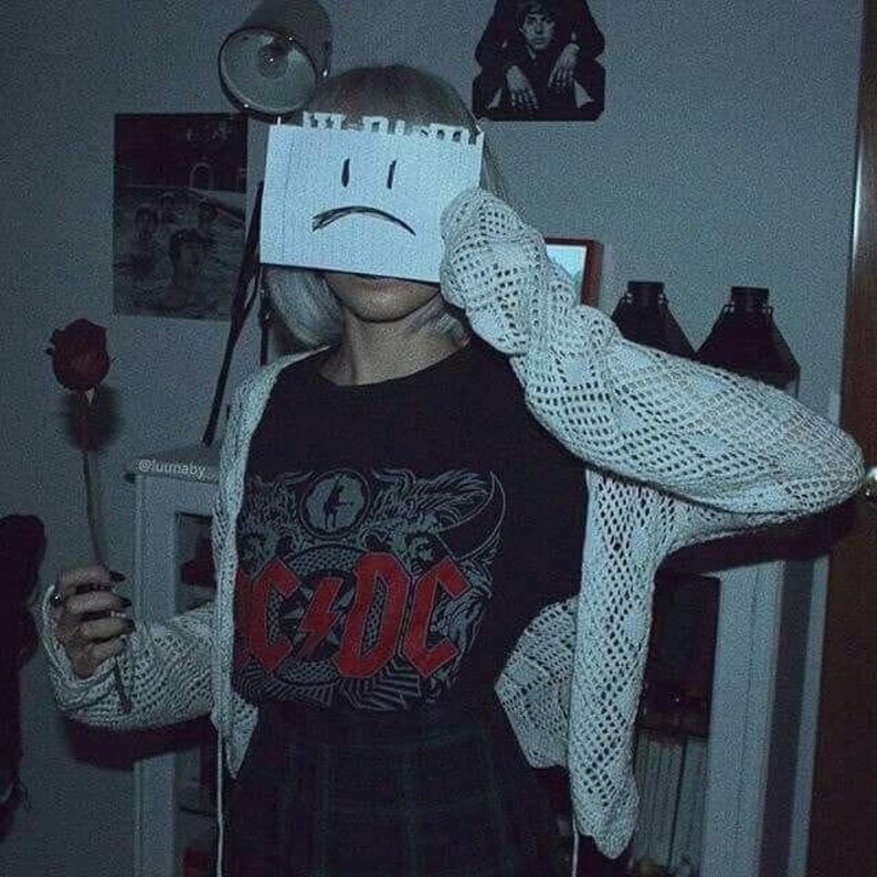 May include: A person wearing a black t-shirt with a red AC/DC logo, a white crocheted cardigan, and a plaid skirt. They are holding a single red rose and covering their face with a piece of paper with a sad face drawn on it.