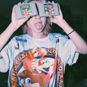 May include: A person wearing a blue and orange t-shirt with the words "Banjo Kazooie" and "Diddy Kong Racing" printed on it. The person is holding two Nintendo 64 game cartridges in front of their face.