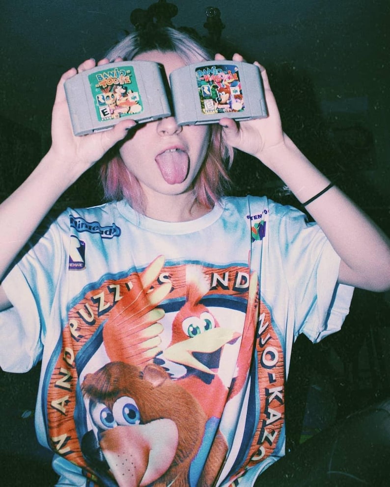May include: A person wearing a blue and orange t-shirt with the words "Banjo Kazooie" and "Diddy Kong Racing" printed on it. The person is holding two Nintendo 64 game cartridges in front of their face.