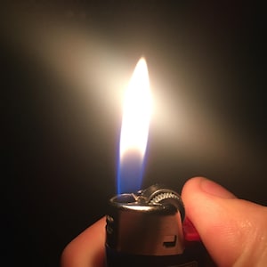 May include: A blue and silver lighter with a red top is lit, showing a bright orange flame. The lighter has the word "BIC" printed on the side.