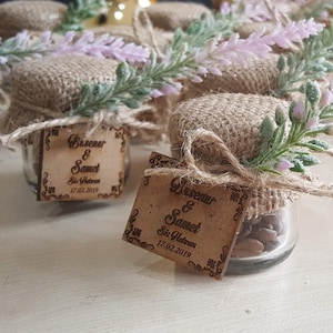 Peut inclure: Petits pots en verre avec des couvercles en toile de jute et une brindille de lavande sont liés avec de la ficelle. Chaque pot a une étiquette en bois avec les noms "Busenur & Samet" et la date "17.02.2019" gravés dessus.