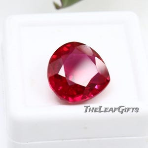 Puede incluir: Una gema de rubí facetada en forma de corazón, que muestra un tono rojo intenso con facetas internas más claras. La gema está sobre un fondo blanco, con el texto "TheLeafGifts" visible.