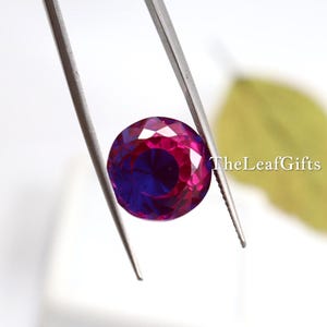 May include: A round, faceted gemstone held by tweezers. The gem has a deep magenta hue on the outer edges, transitioning to a rich, dark blue in the centre. TheLeafGifts is printed on the right side.