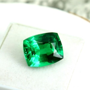 May include: A vibrant green emerald gemstone with a cushion cut, displayed on a white background.