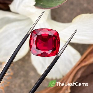 Op de afbeelding: Een levendige, kussenvormige rode edelsteen wordt door een pincet vastgehouden tegen een vage achtergrond van witte bloemblaadjes en bladeren. De facetten van de edelsteen reflecteren licht. Het The Leaf Gems-logo is zichtbaar.