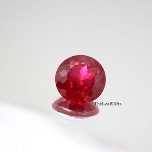 Könnte beinhalten: Ein facettierter, runder, tiefroter Edelstein mit Brillantschliff. Der Edelstein ist hochreflektierend, mit einem hellrosa Zentrum und einer Spiegelreflexion darunter. TheLeafGifts ist rechts aufgedruckt.