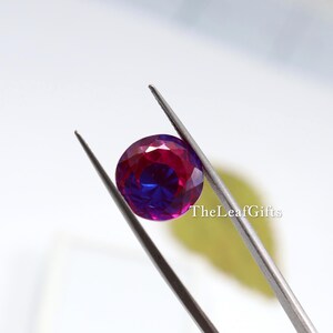 May include: A round, faceted gemstone held by tweezers. The gem displays a colour gradient, shifting from deep red to vibrant blue. TheLeafGifts is printed on the right side of the image.