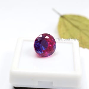May include: A round, faceted gemstone displaying a mix of deep purple and magenta colours. The gem is presented in a white display case. TheLeafGifts is printed on the right side of the image.