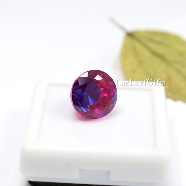 May include: A round, faceted gemstone displaying a mix of deep purple and magenta colours. The gem is presented in a white display case. TheLeafGifts is printed on the right side of the image.