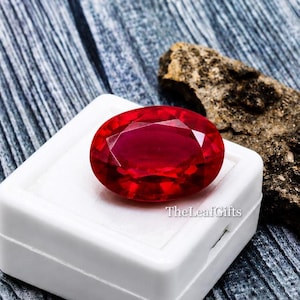 Può includere: Un'intensa gemma di rubino taglio ovale, esposta in una scatola bianca. Il rubino è di un colore rosso intenso con sfaccettature che catturano la luce. Lo sfondo presenta una superficie scura e strutturata e un pezzo di pietra grezza. TheLeafGifts è stampato.