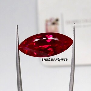 May include: A vibrant red marquise-cut gemstone held by silver-toned metal tweezers. The faceted stone displays a brilliant crimson hue with light reflections across its surface. The text THELEAFGIFTS is printed in black below the gemstone.