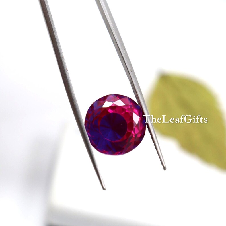 May include: A round, faceted gemstone held by tweezers. The gem has a deep magenta hue with a hint of purple in the centre. TheLeafGifts is printed on the right side of the image.