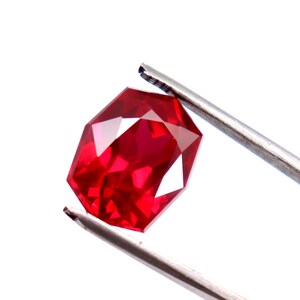 May include: A deep red gemstone with a faceted cut, held by a pair of tweezers.