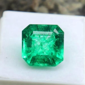 Peut inclure: Une émeraude verte vibrante de forme rectangulaire avec une apparence légèrement nuageuse. L'émeraude est présentée sur une surface blanche.
