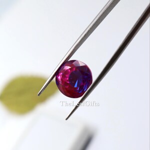 May include: A round, faceted gemstone, displaying a mix of pink and blue colours, is held by tweezers. The gem's surface reflects light, highlighting its cut. The background is blurred, with a leaf and the text "TheLeafGifts".