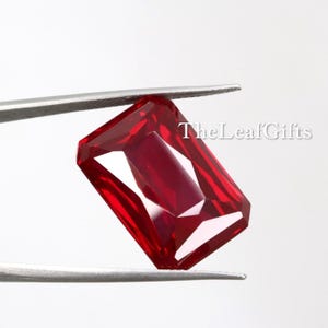 Può includere: Una gemma rossa dal taglio rettangolare tenuta da una pinzetta metallica color argento. La pietra presenta una superficie sfaccettata che riflette la luce, creando profonde tonalità cremisi e riflessi luminosi. Il testo TheLeafGifts è visibile in bianco.