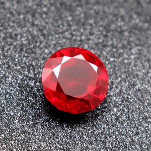 Puede incluir: Una piedra preciosa de color rojo intenso con un corte facetado. La piedra preciosa tiene forma ovalada y un alto pulido.