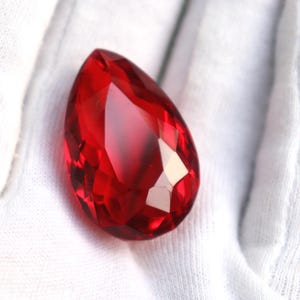 May include: A faceted, teardrop-shaped, red gemstone. The gem is a deep, rich red color and has many reflective surfaces. The gem is resting on a white, textured surface.