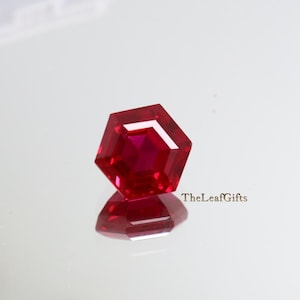 Könnte beinhalten: Ein facettierter, sechseckiger Rubin-Edelstein in tiefroter Farbe. Der Edelstein wird auf einer reflektierenden Oberfläche präsentiert, die seine Facetten und Brillanz zeigt. TheLeafGifts ist auf der rechten Seite aufgedruckt.
